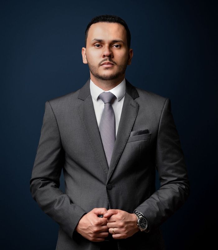 Advogado criminalista sério em pé com braços cruzados, terno preto, fundo neutro escuro, retrato profissional de estúdio, iluminação de contraste
